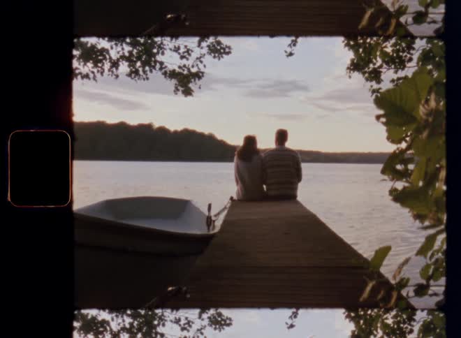 A man and woman sitting on a dock next to a boat overlooking a lake of water during daytime while dating on a warm summer day on Super-8 mm film