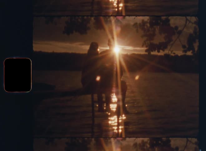 A young man and woman sit on a dock over water watching the sunset and the reflection on the lake as a light breeze makes the branches of the trees move and the surface ripple on old Super 8mm film