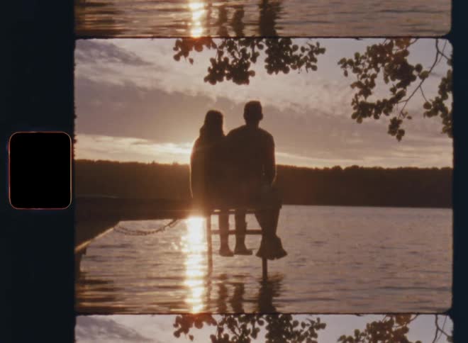 Panning 8mm shot of a young loving couple watching a lakeside sunset from a dock during a romantic date with backlight and bright lens flares