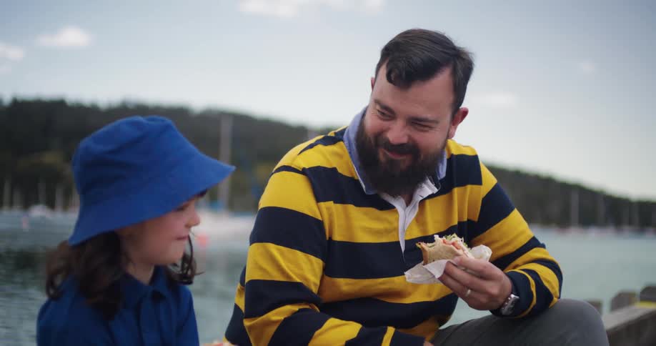 A father sits on the shore with his daughter and laughs as they talk and eat sandwiches together