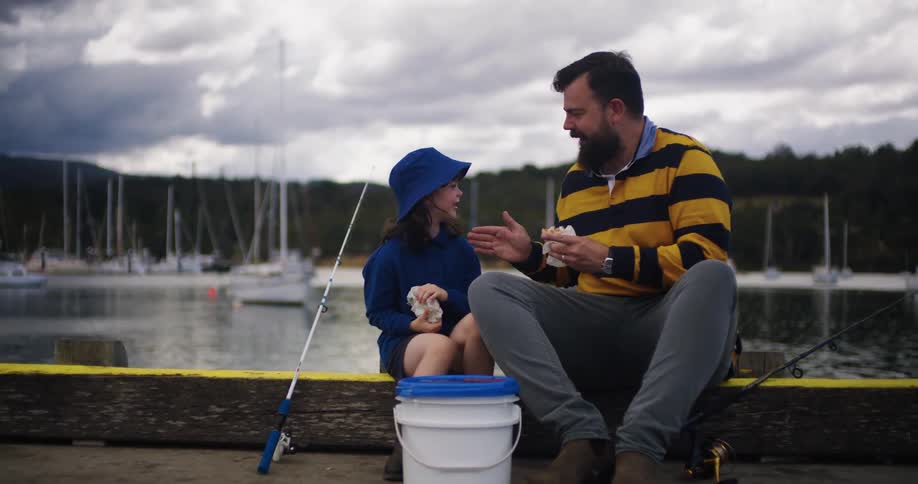 A man is sitting along the dock with his young daughter eating lunch with a fishing pole and a bucket nearby