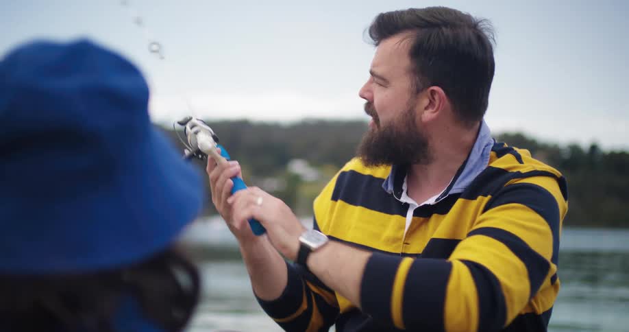 A man in a striped shirt holds a fishing pole while explaining how it works to a person wearing a blue hat