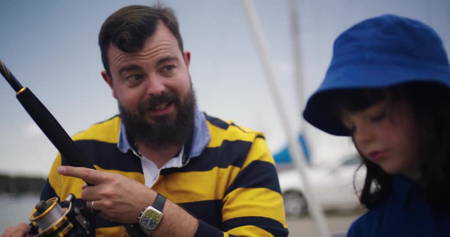 Father with beard black and yellow striped shirt and wristwatch and girl with dark wavy hair and blue hat and shirt fish on the ocean shore