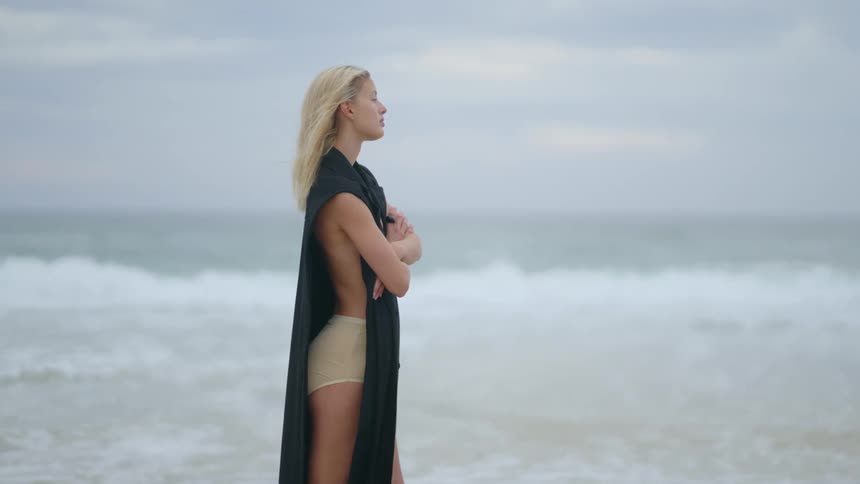 A scantily clad woman walks on the shore and then stops and crosses her arms over her chest