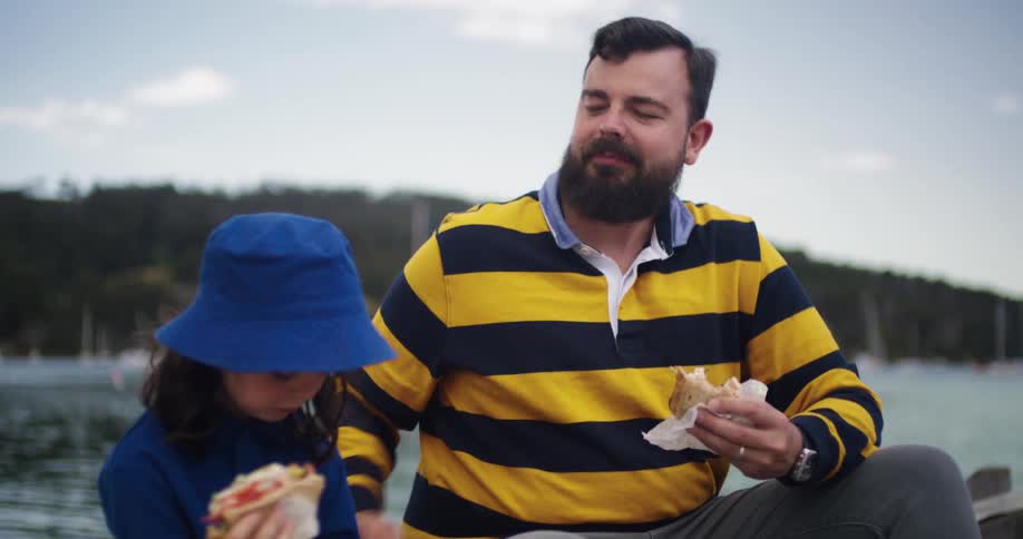 Father with black and yellow sweatshirt and gray pants eats sandwiches with daughter wearing blue hat and shirt on the coast