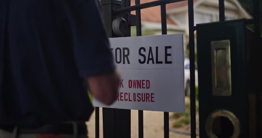 Man hangs for sale sign of bank owned foreclosure on gate of large house with car sitting in driveway