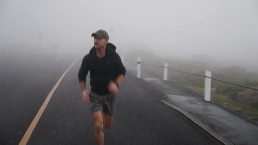 Man with cap black hoodie and shorts jogs on highway in fog