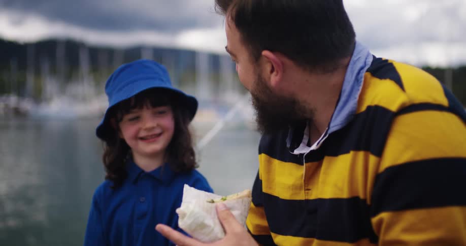 A man and his daughter eat sandwiches while sitting on a pier