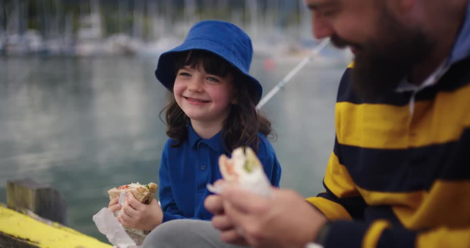 A father sits on a dock with his daughter and they talk while eating sandwiches