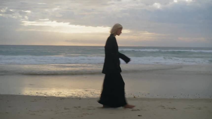 Woman walks along beach wearing long skirt as surf rolls in