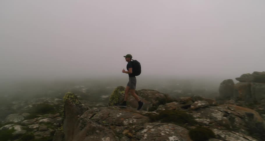 Man with cap shirt shorts and backpack runs in rocky field in fog