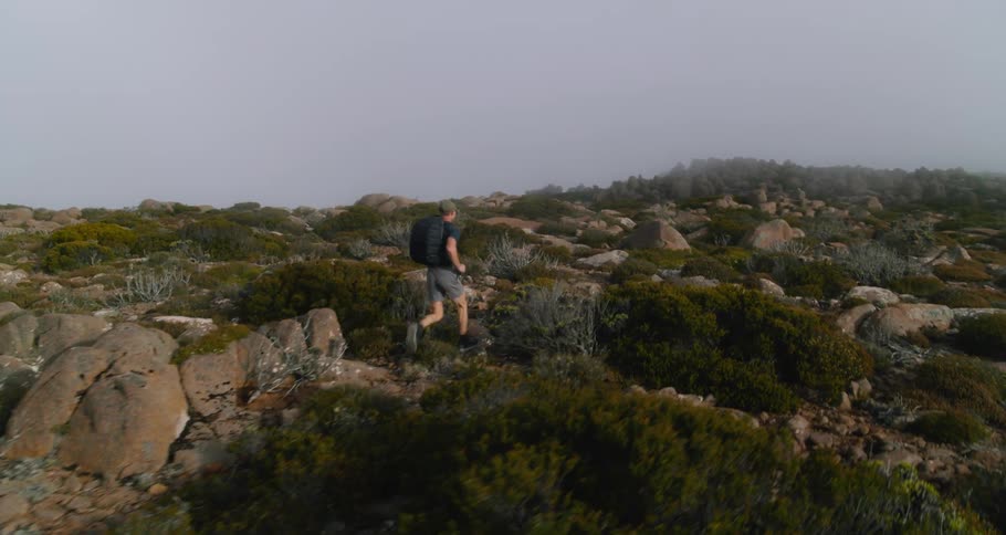 A man wearing a backpack is running across rough terrain covered with rocks and boulders