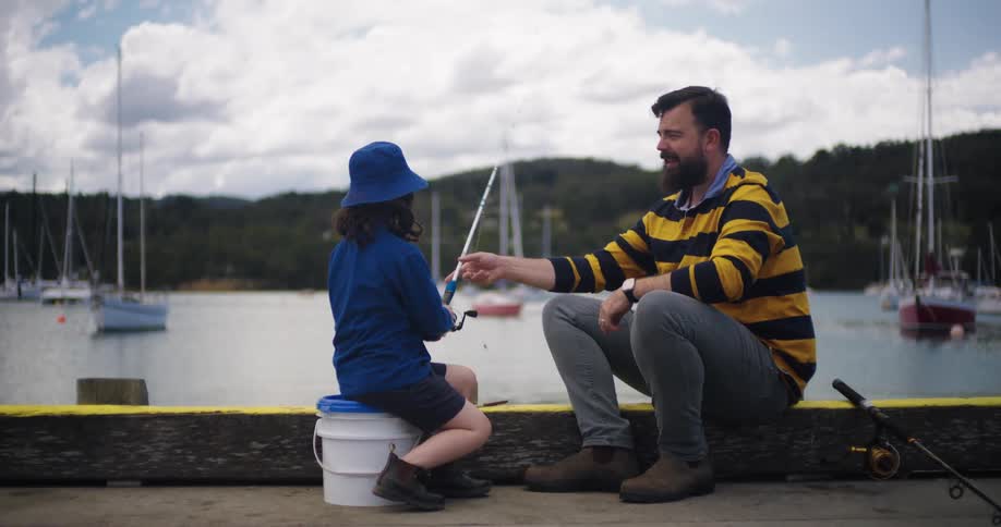 A little girl is on the dock with her father and he hands her the fishing pole as she sits on a bucket