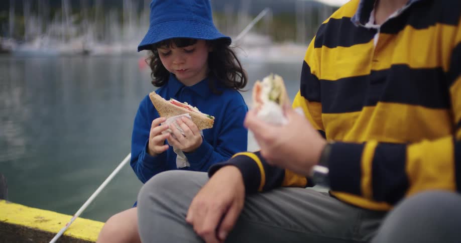 Father and daughter eat sandwiches while fishing across from marina