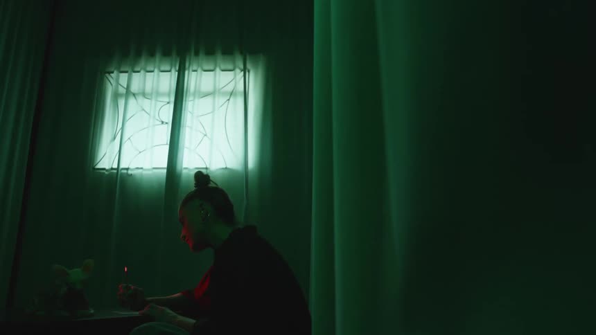 Light shines in through the curtains as a woman sits in a dark room with a stuffed animal on the table as she writes on paper