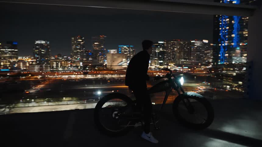 A man riding a motorbike on a rooftop at night gets off and stands at the railing overlooking a city
