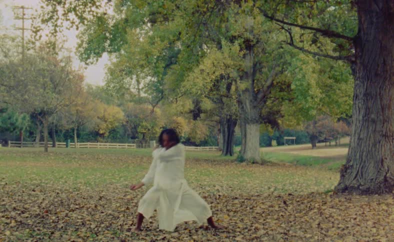 A woman wearing a white outfit is outside in her leaf covered yard waving her arms and dancing near a tree
