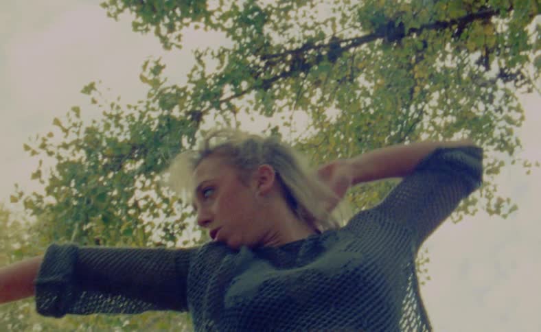 A woman is standing outside under a tree moving her arms and head as she dances in a dramatic way
