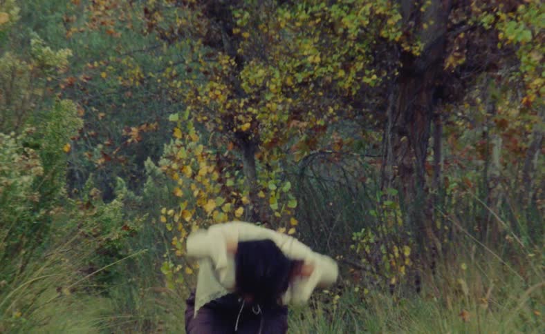 A teenage girl is standing near trees in the woods moving her arms and head as she dances