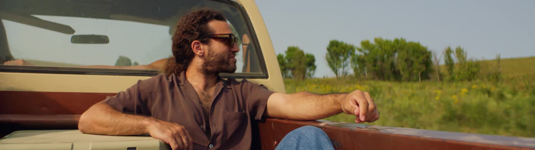 A man is sitting in a truck bed while two people are in the cab as they travel in a rural area with grassy fields and trees in the daytime