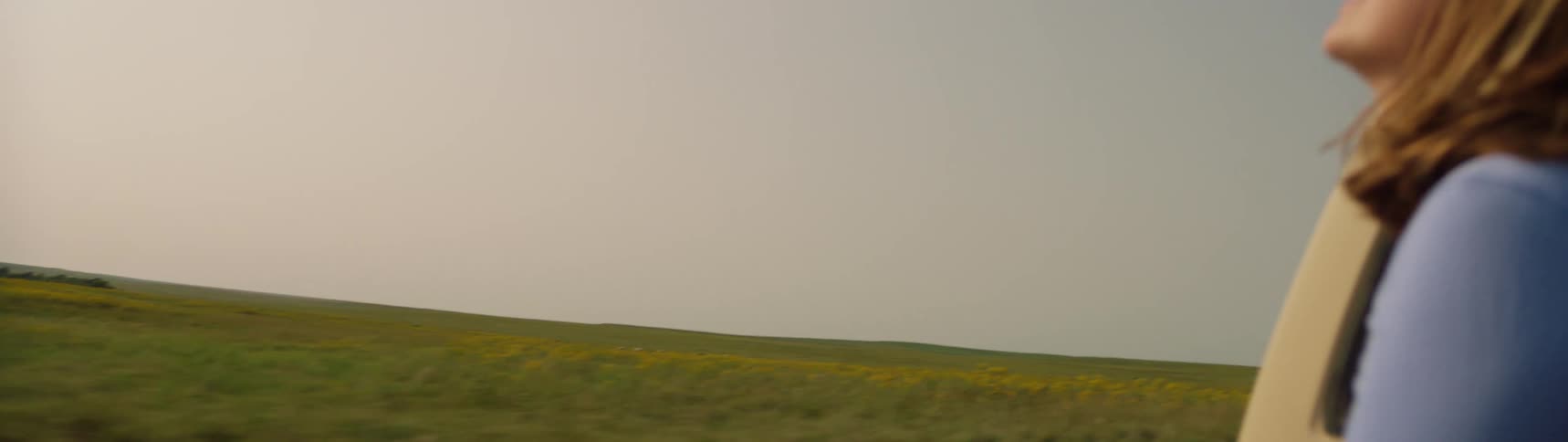A woman is sitting in a pickup truck bed as it travels by a large rural field of grass in the evening