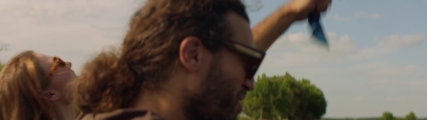 Man with sunglasses and curly hair brown t shirt sits next to woman with white shirt dancing near the lake in the field