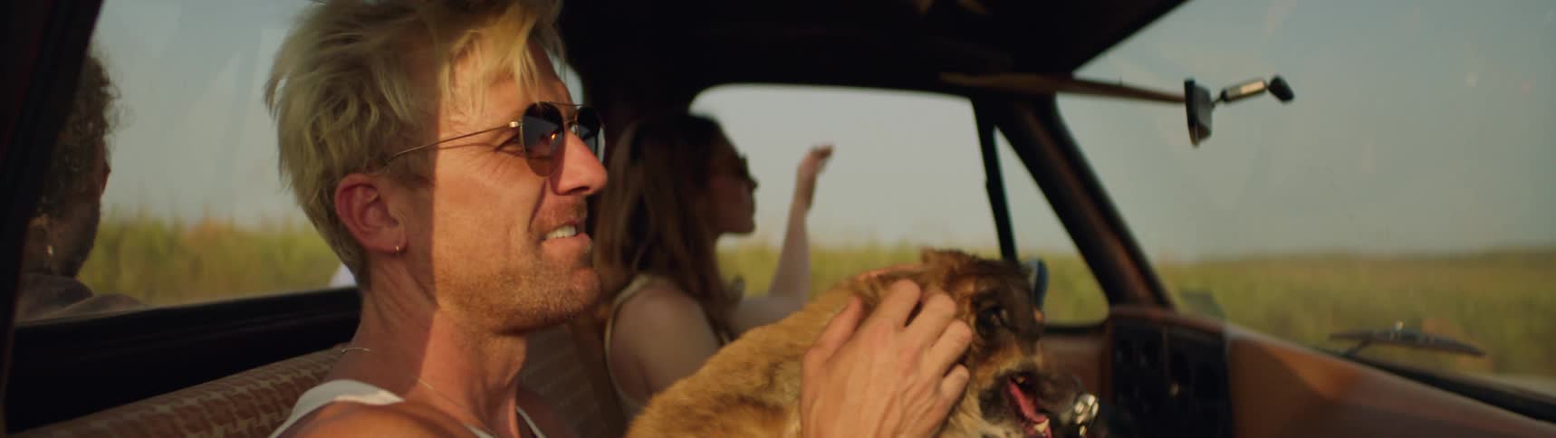 Man pets dog and rides in truck cab and another passenger sits in truck bed as woman drives truck past field