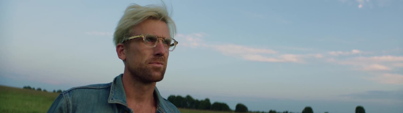 Man with blonde hair putting on glasses while standing in a pasture