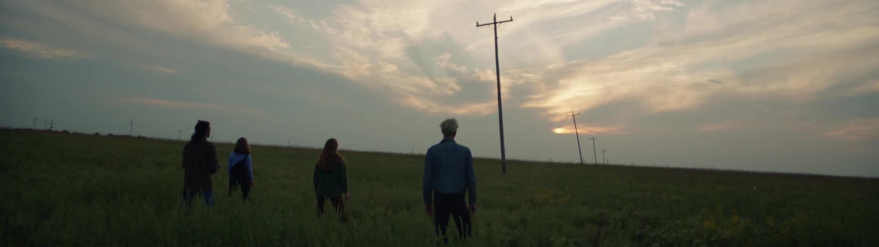 Four people standing in a grassy field along a line of telephone poles at dusk