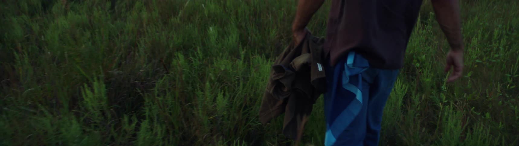 A man walks through a vast field while carrying a jacket during the day