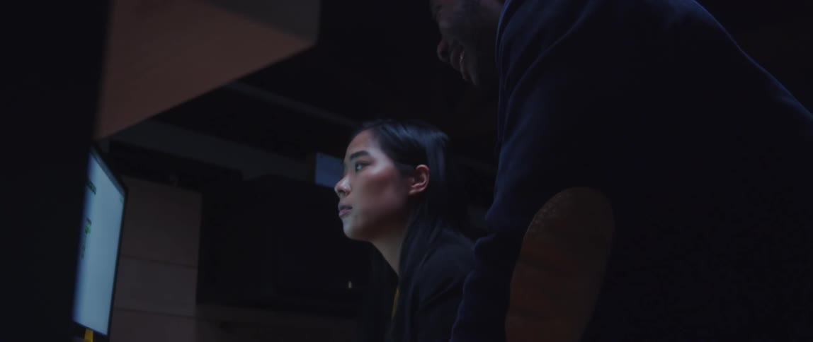 A woman sits in front of a computer at an office and listens to a man as he talks and points at the screen