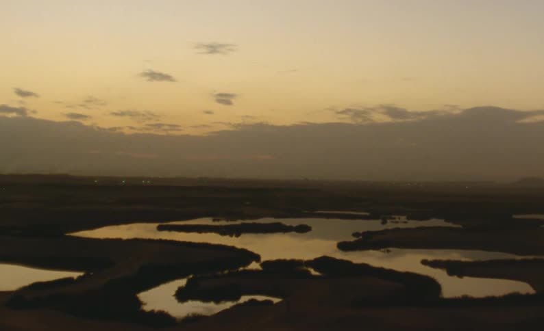 Winding water reflects red tinted sky with electric lights near the horizon