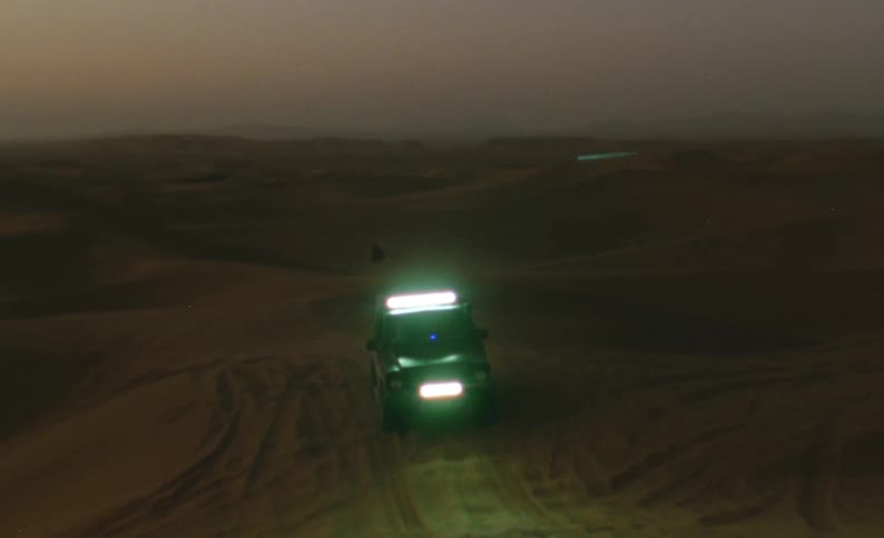 A jeep with headlights on driving through the desert at dusk