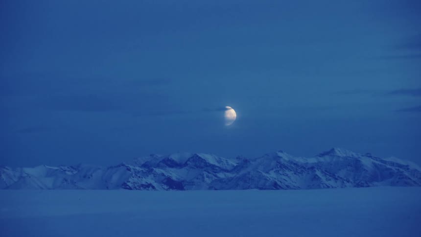 The moon shines brightly above a snow covered mountain range as the sky grows darker