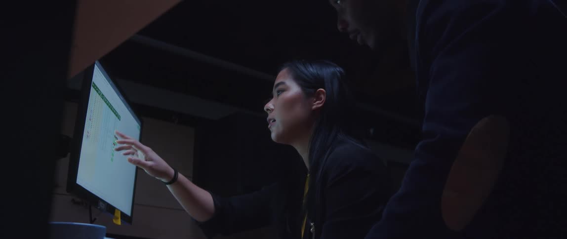 A woman is sitting at a desk as she points to a computer monitor and talks to a man looking over her shoulder