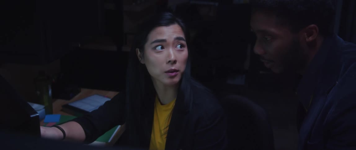 A woman is sitting at a desk as she points to a computer monitor and talks to a man looking over her shoulder