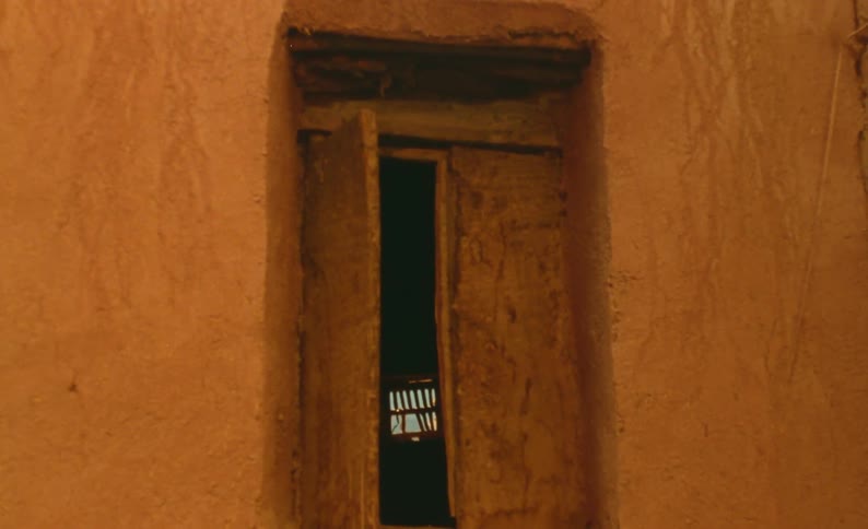The door to an adobe building is open and a light from a window shines in the darkness