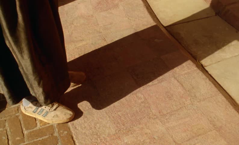A person wearing white and blue tennis shoes and dark pants is standing on a brick road and casting a shadow across the walkway