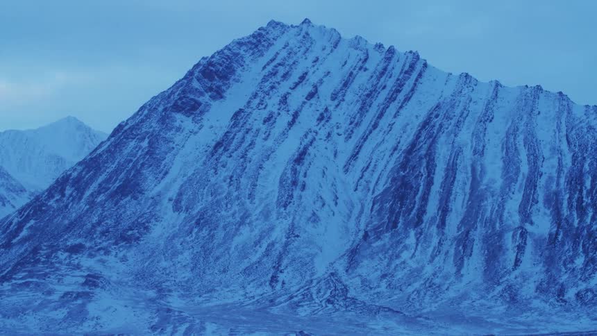 A snow covered mountain peak with ridges down its side and mountains in the background