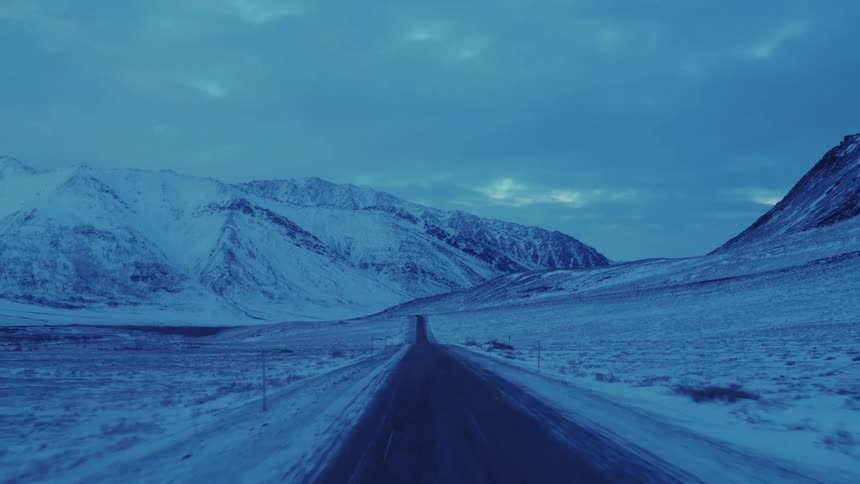 A long paved road with snow covered roadsides and mountains in the distance