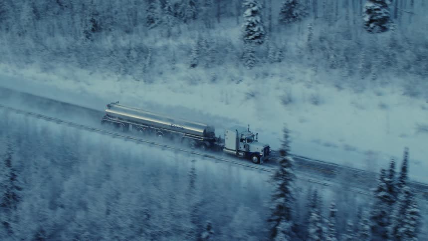 A tractor trailer driving down a road in winter with snow covered grounds and trees around the road