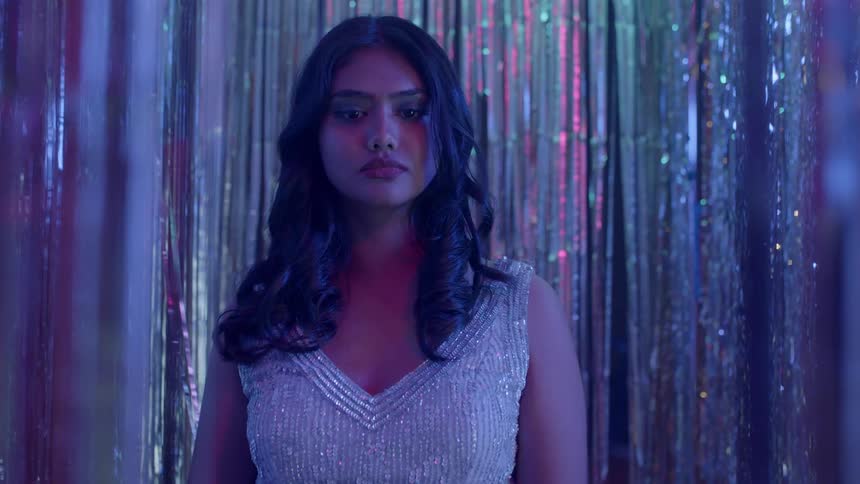A woman with long dark hair looks nervous as she stand on a stage as lights reflect on the shiny tinsel behind her