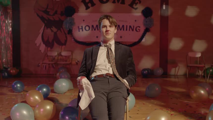 A young man sits on a chair at a school dance with a bloody nose