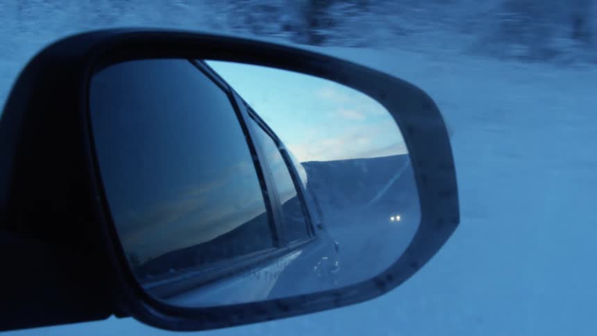 A side mirror on a car viewing the car behind it on the snow covered road