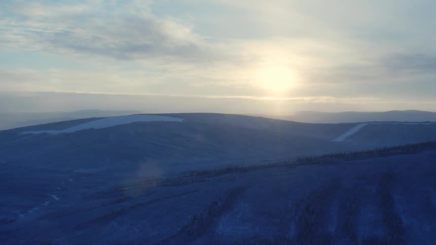 A snowy hillside with pine trees and the sun setting over the hills