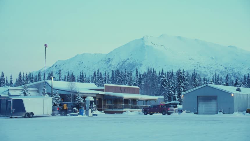 Two people are standing next to a truck outside of buildings at the foot of a mountain with a forest on a snowy winter day