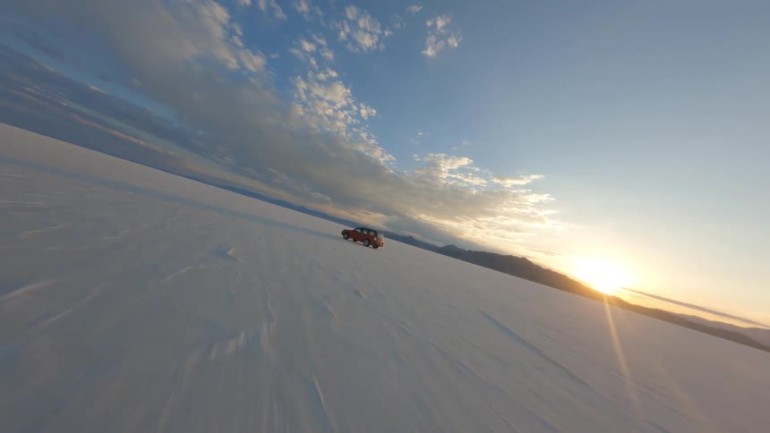 Red vehicle drives across snow covered plain as sun rises over mountains
