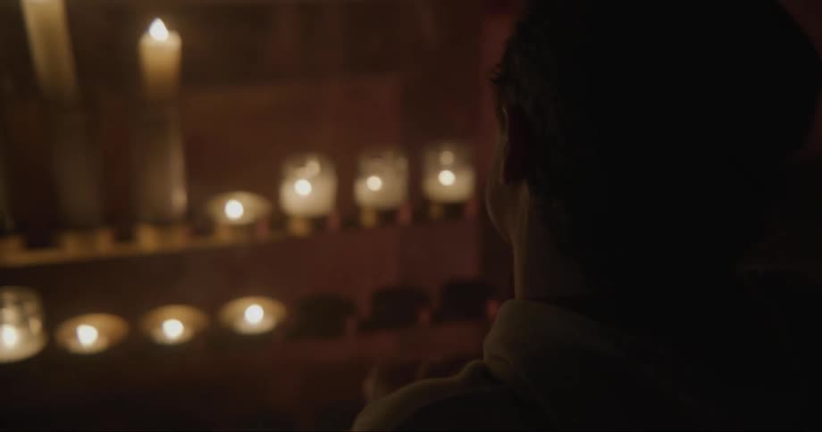 Young man kneels in prayer in front of a row of candles at night