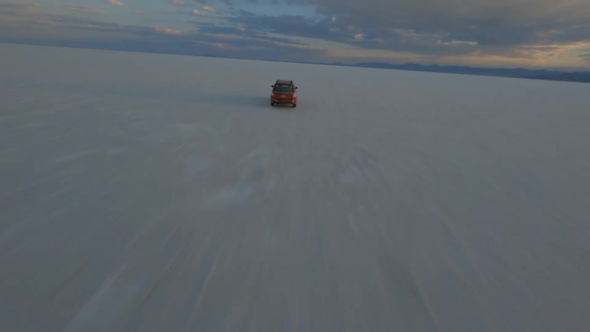 Red vehicle drives across snow covered plain toward mountains under a cloudy sky