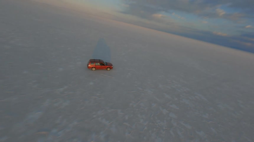 Red vehicle drives across snow covered plain as sun sits behind mountains on horizon